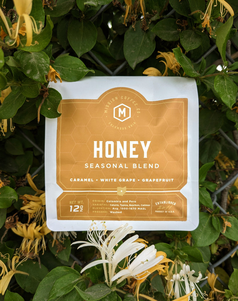 Locally roasted coffee from Columbus Ohio, ethically sourced, sustainable practices.  Yellow Orange bag set against chain link overgrown with honeysuckle vines.